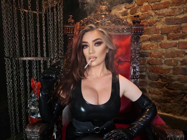 MiaBlackXO with big tits, wearing black leather gloves and dress, sitting on an ornate chair, smoking a cigarette, with dark makeup and long brown hair.
