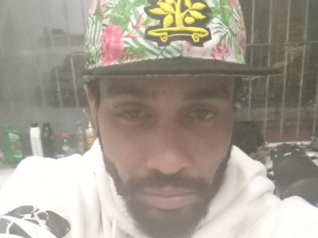 GabiZera has a dark brown complexion, short beard, and is wearing a floral-patterned cap with a yellow emblem. He's in a white hoodie, looking serious.