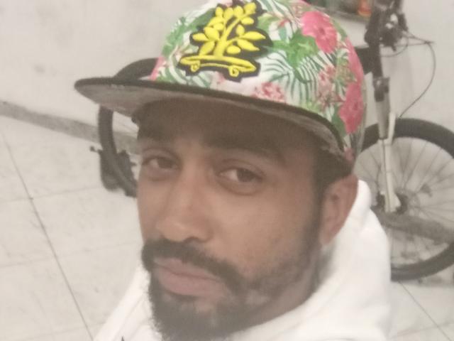 GabiZera with a floral and pink baseball cap, black beard, and white hoodie, looking slightly bored. Bicycle in the background. Soft-focus image.