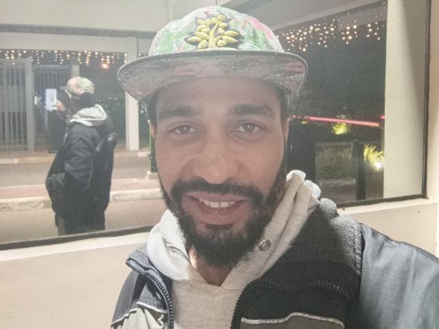GabiZera, usando um chapéu floral e uma jaqueta preta com moletom branco, tem barba e sorri para a câmera. Seu reflexo é visível na janela atrás dele..