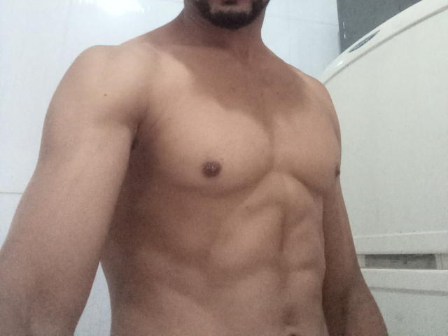 GabiZera's bare, muscular chest with small, dark nipples and defined abs, standing in a bathroom, only showing from neck to waist. No visible genitals. Bearded, medium skin tone.