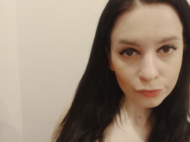 ScarlettAmore a la peau pâle, un trait d'eye-liner noir et de longs cheveux noirs. Elle regarde l'appareil photo avec un léger sourire, les lèvres légèrement pincées. Ses petites seins sont à peine visibles..