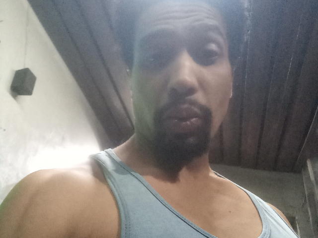 GabiZera with a light brown skin tone, short beard, and mustache, wearing a light blue tank top, looking into the camera with a slightly confused expression.
