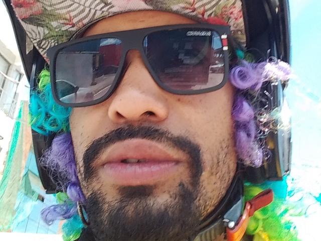 GabiZera with black sunglasses, curly purple and green hair, beard, and helmet, reflecting a building in their visor. GabiZera's nose and lips are centered in the shot.