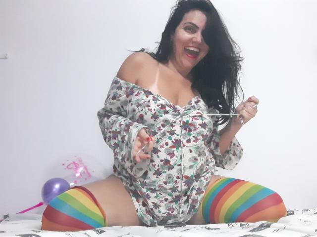 PammyBurnher with long black hair, smiling, wearing a floral off-shoulder top and rainbow-striped thigh-highs, sitting on a bed with her tits and ass visible.