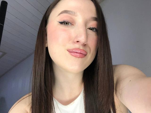 MonAmourR a les lèvres roses et pulpeuses, les cheveux bruns et lisses, et un trait d'eye-liner ailé, elle tire légèrement la langue avec un petit sourire. Elle porte un débardeur blanc..