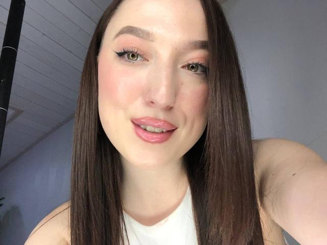 A MonAmourR tem pele clara, olhos verdes e lábios rosados, com cabelos castanhos longos e delineador preto, sorrindo levemente, vestindo uma blusa branca, em um selfie..