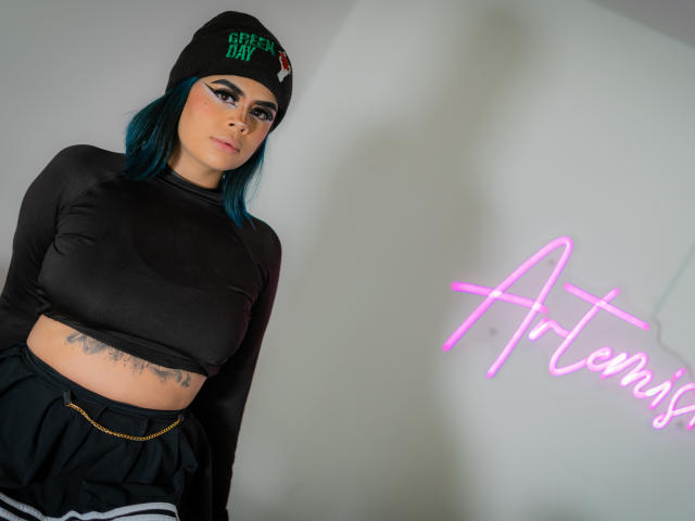 MissFontainne with blue hair, black beanie, and "Green Day" text, wearing a black long-sleeve crop top, black skirt, and showing tattoos on her belly. Neon sign reads "Artexis."