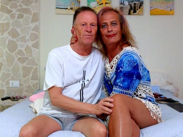 GiginDave and GiginDave's wife sitting on a bed, GiginDave in a white tee and shorts, wife in a blue and white patterned dress, both smiling.