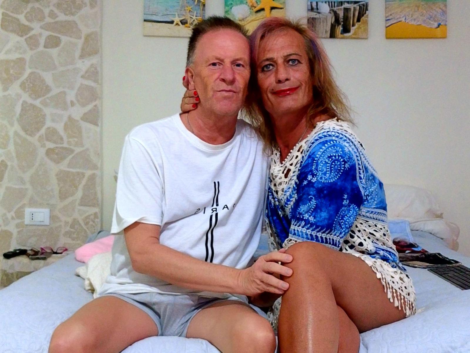 GiginDave and GiginDave's wife sitting on a bed, GiginDave in a white tee and shorts, wife in a blue and white patterned dress, both smiling.