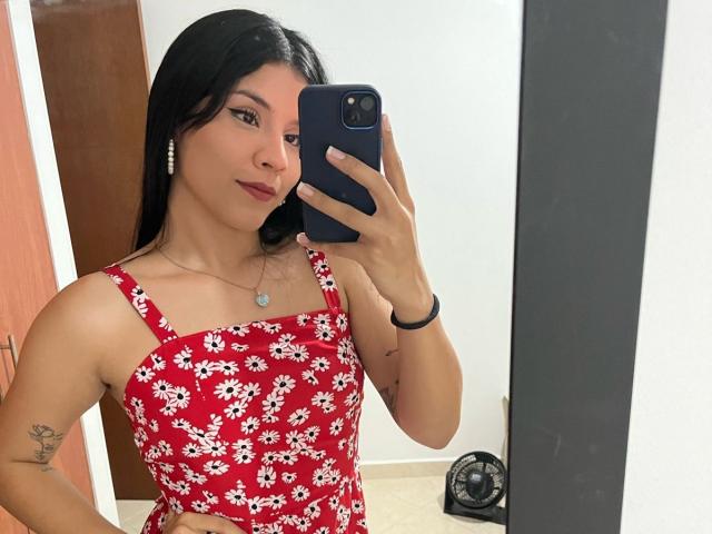 KatyGreyy with black hair, red floral dress, holding phone, small tattoo on arm, wearing necklace, standing in front of mirror, smiling slightly, phone in right hand.