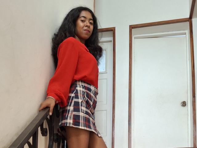 LaeticiaPink with long black hair, red long-sleeve shirt, and plaid skirt, standing on stairs, hand on rail, looking slightly to the side, with a confident expression.