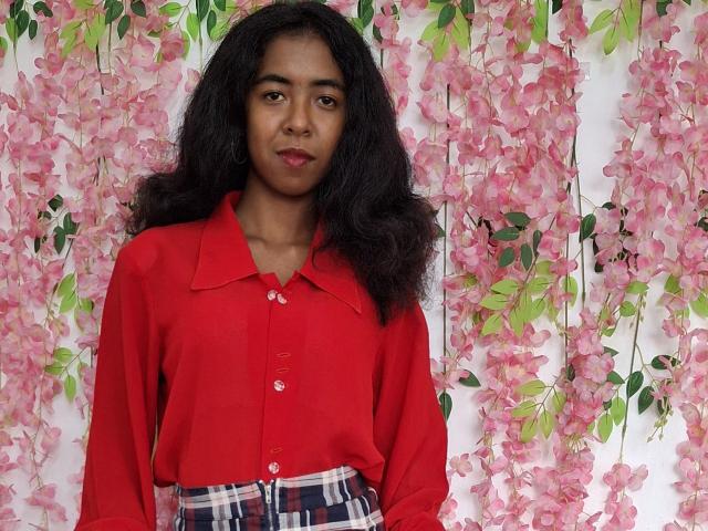 LaeticiaPink with medium brown skin, wavy black hair, red button-up shirt, plaid skirt, standing against pink flower wall, looking slightly to the side, red lipstick.