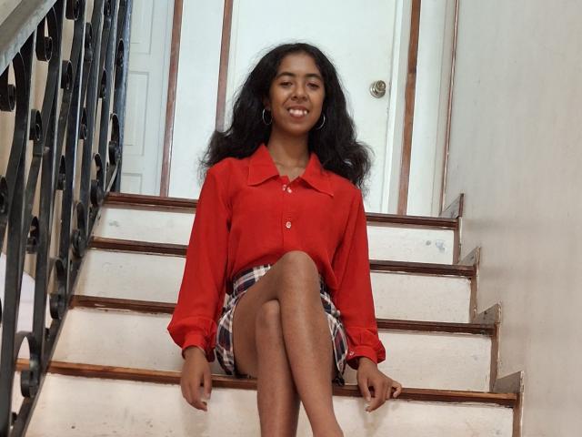 LaeticiaPink with dark curly hair, wearing a red shirt and plaid skirt, sitting on white stairs with legs crossed, smiling, and wearing hoop earrings.