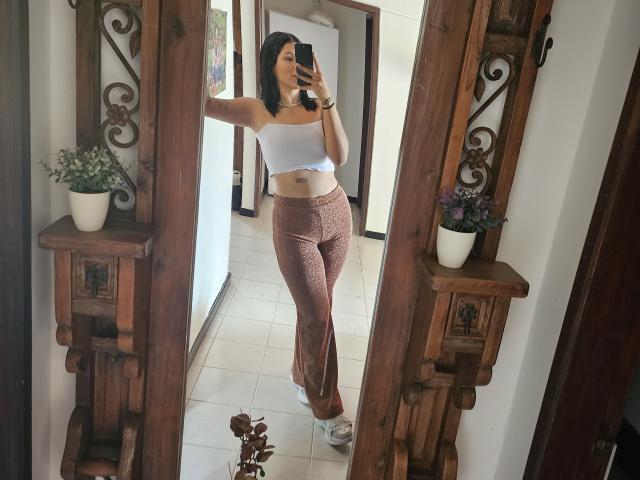KatyGreyy with small tits, wearing a white crop top and pink patterned pants, taking a mirror selfie, holding a phone, standing on white tile floor, wooden mirror frames.