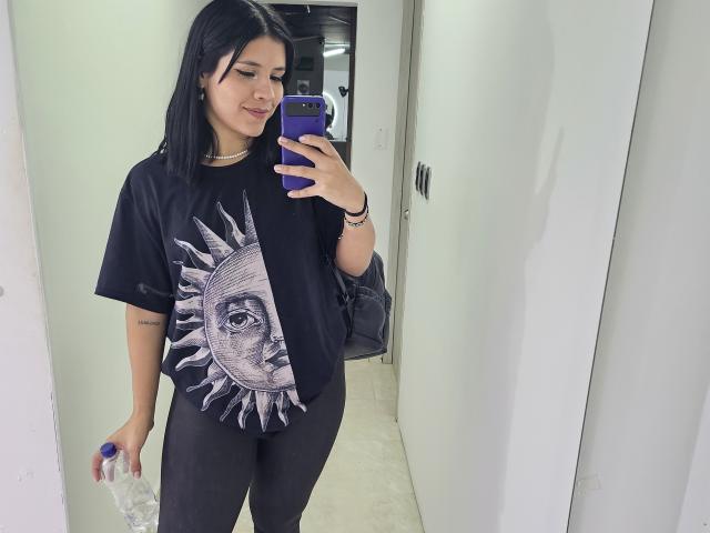 KatyGreyy with black hair, wearing a sun graphic tee, black pants, holding a phone with a purple case, smiling, and holding a water bottle.