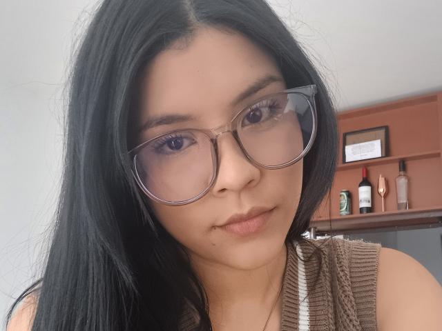 KatyGreyy with long black hair, wearing large glasses, brown sweater, and subtle makeup, looking directly at the camera with a slight smile. Her pussy is not visible.
