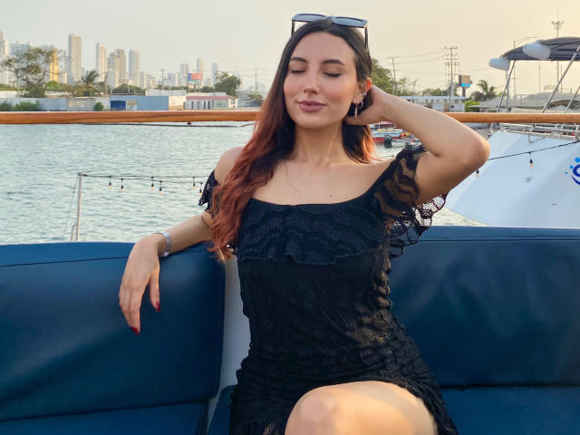 CarlaFranco met lang rood haar, gekleed in een zwarte kanten jurk, zittend op een blauwe boot, hand in haar haar, zonnebril op haar hoofd, ogen gesloten, subtiele glimlach, water en stad op de achtergrond..