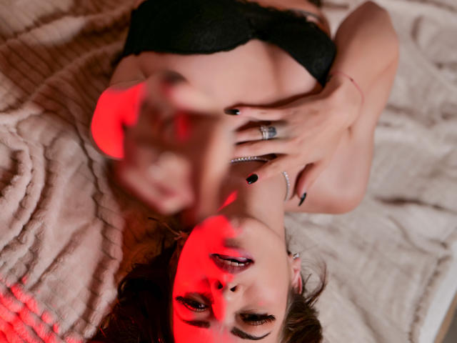 RinaQueenie lying on a bed, wearing black lingerie, with red light on her face, hand on her chest, tongue out, black nails, and a ring on her finger.