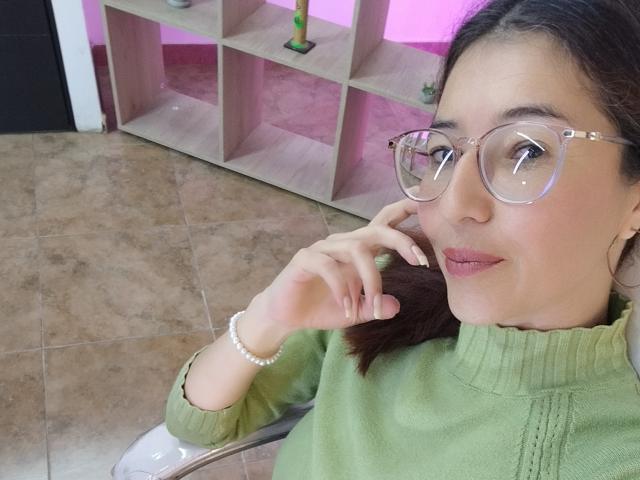 AntonnellaS with glasses, green turtleneck, and pearl bracelet, touching her cheek, brown hair, pink light, tiled floor, and wooden shelf in background. Her ass is not visible.