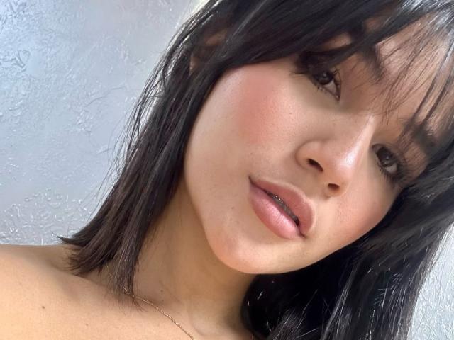 ScarlletZhen's face, showing her smooth, fair skin with a slight blush, black straight hair with bangs, and full pink lips, looking slightly to the side with a subtle smile.