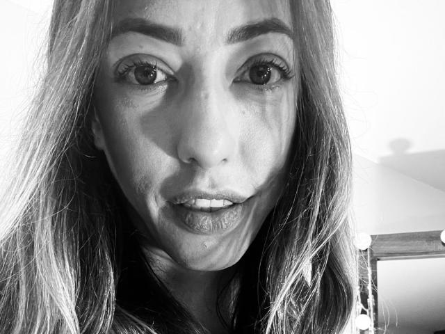 Black and white selfie of HollyPinky with long hair, thick eyebrows, and slightly parted lips showing teeth. Her eyes are wide and she has a small nose and lips.