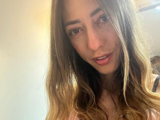 HollyPinky ha lunghi capelli castani, pelle chiara e un trucco leggero con rossetto rosa. Le sue sopracciglia sono leggermente alzate e guarda la fotocamera con un sorriso dolce..