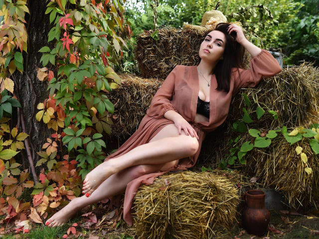 WilsonMelanie with pale skin, dark hair, and red nails, wearing a brown dress, sits on hay, legs crossed, hand in hair, showing her bare ass, next to a ceramic pot.