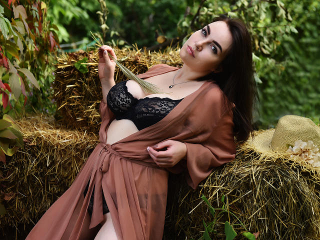 WilsonMelanie with brown eyes, dark hair, wearing a black lace bra and open brown robe, holding straw, leaning on hay, looking at camera, with a tan hat beside her.