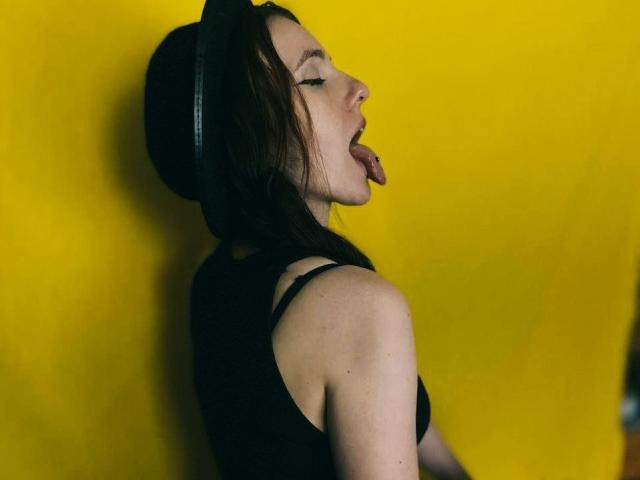 KatKitty with tongue out, wearing a black tank top and headphones, against a yellow wall, showing her bare ass slightly from the side. Her eyes are closed.