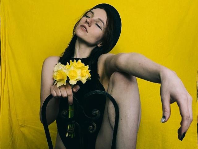 KatKitty with pale skin, dark hair, black headscarf, holding yellow flowers, sitting on black chair, eyes closed, arm stretched out, showing bare ass, against bright yellow backdrop.