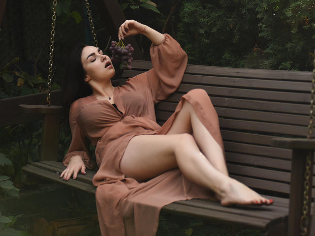 WilsonMelanie is laying on a wooden bench, wearing a sheer brown dress, holding grapes to her mouth, with her legs crossed, showing her bare ass and red-painted toenails.