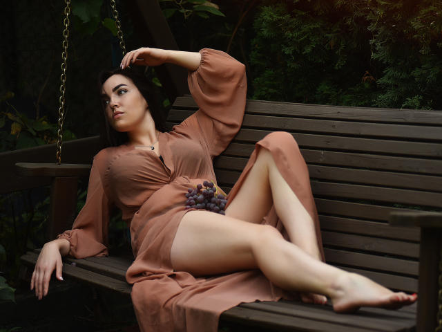 WilsonMelanie, with her ass and pussy partially visible, lounges on a wooden bench, wearing a brown dress, legs spread, grapes in hand, dark hair, looking to the side.