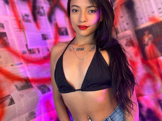 Maite, con lunghi capelli neri, rossetto rosso, reggiseno nero che mette in mostra il seno, tatuaggio "1998" sul petto, collana e piercing all'ombelico, in piedi con i jeans blu..