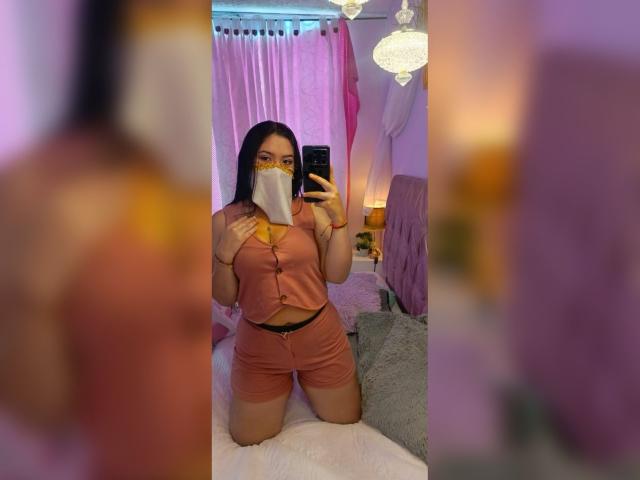 AlanaHalamm, con cabello oscuro, usando una mascarilla facial, una blusa rosa abotonada y pantalones cortos a juego, está arrodillada en una cama, sosteniendo un teléfono, mostrando sus curvas y sus pequeños senos..