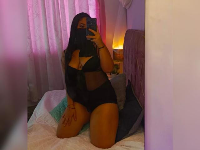AlanaHalamm con lunghi capelli neri, che indossa lingerie nera, inginocchiata sul letto, selfie allo specchio, con il telefono in mano, il viso coperto dai capelli, un tatuaggio visibile sulla coscia destra, letto con illuminazione rosa e viola..