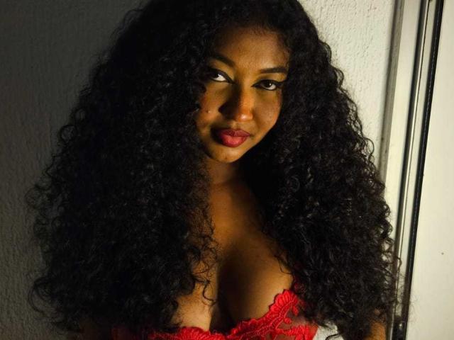 ValeriSagem has dark curly hair, red lipstick, and a seductive look, wearing a red lace bra that accentuates her large, curvy tits. She's standing in dim light.