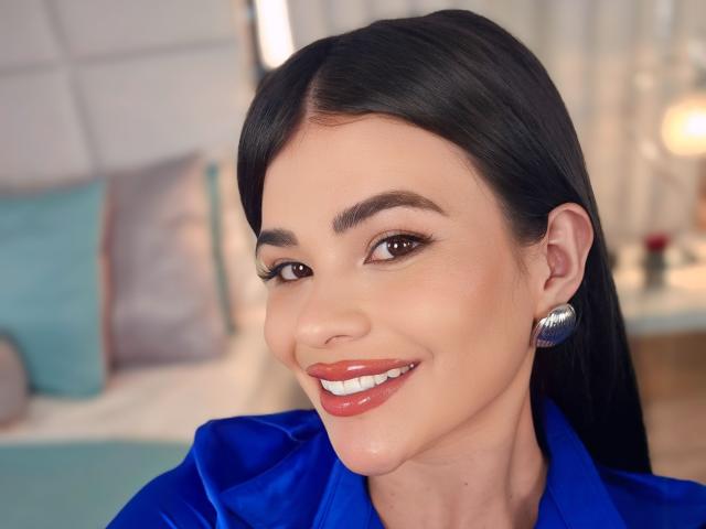 PaulinaValley with shiny red lipstick, blue satin shirt, and silver earrings, smiling at the camera with brown eyes and straight black hair. Her tits are not visible.
