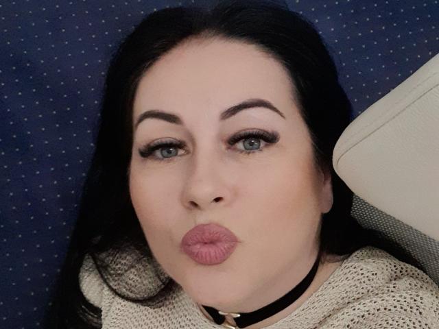 KyraK with pouty pink lips, blue eyes, and dark eyebrows, wearing a black choker and beige knit sweater, making a kissy face.