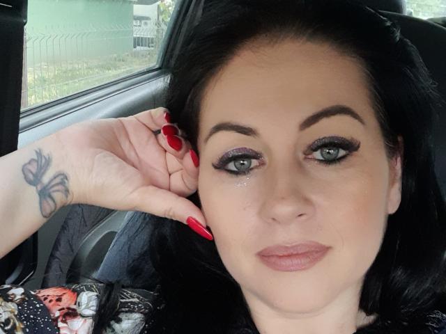 KyraK with black hair, red nails, butterfly tattoo on hand, wearing makeup, green eyes, slight smile, inside car, hand near ear, looking at camera, no visible clothes.