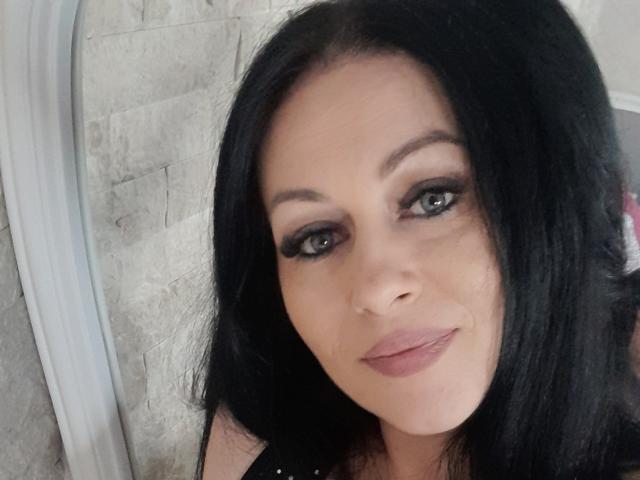 KyraK has dark black hair, green eyes, and wears black eyeliner and pink lipstick. Her face is slightly angled, showing a subtle smile. Her skin is fair.