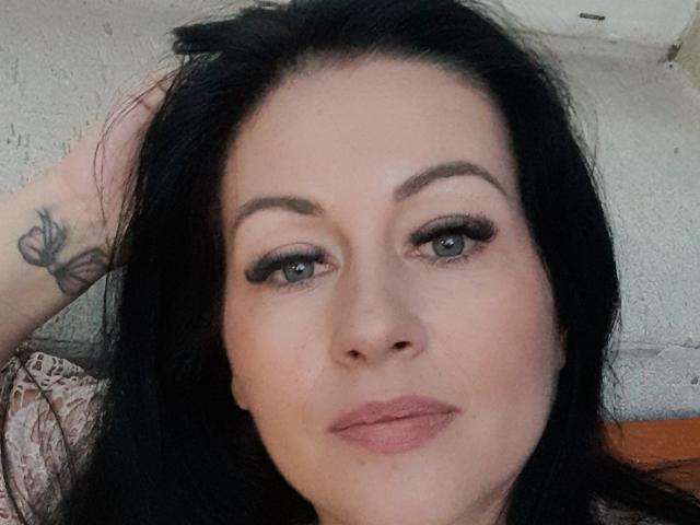 KyraK has black hair, green eyes, and a butterfly tattoo on her left arm. She's wearing black eyeliner and pink lipstick, with a slight smile.