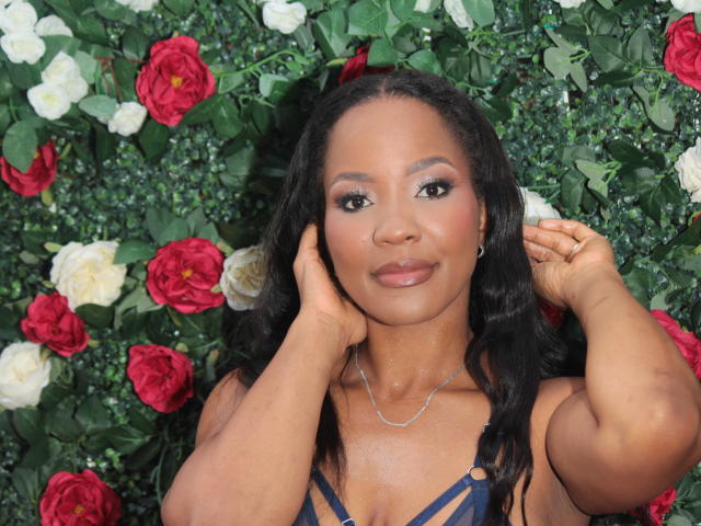 CarmelRoa with dark skin, black wavy hair, and a blue strappy top, hands on her cheeks, smiling, red and white roses in the background, wearing a silver necklace.