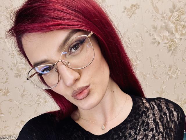 Rosalyn with red hair, wearing glasses, has a serious face, full lips, and is dressed in a black lace top. She's looking at the camera, with a subtle necklace.