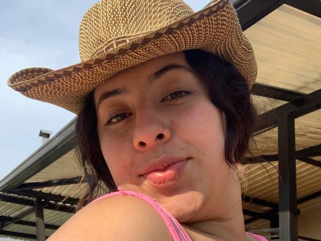 ScarlettParchMusa, wearing a straw hat, has puffy pink lips, a nose ring, and dark hair, with a pink tank top, looking slightly smug in a sunlit shot.