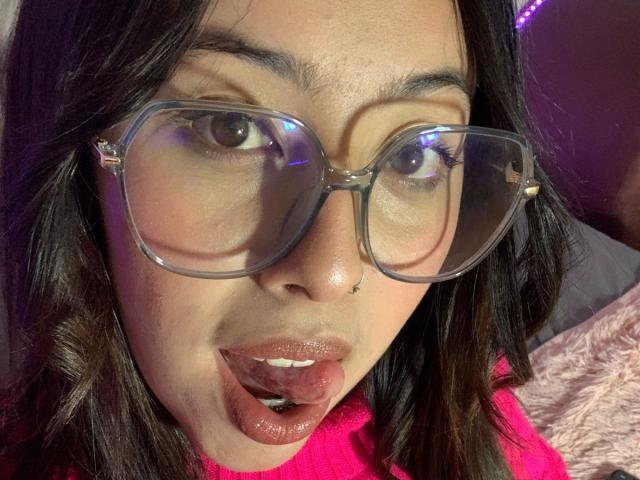 ScarlettParchMusa with glasses, brown eyes, and slightly parted pink lips, tongue touching upper lip, wearing pink shirt, dark hair, nose piercing, soft skin, looking up.