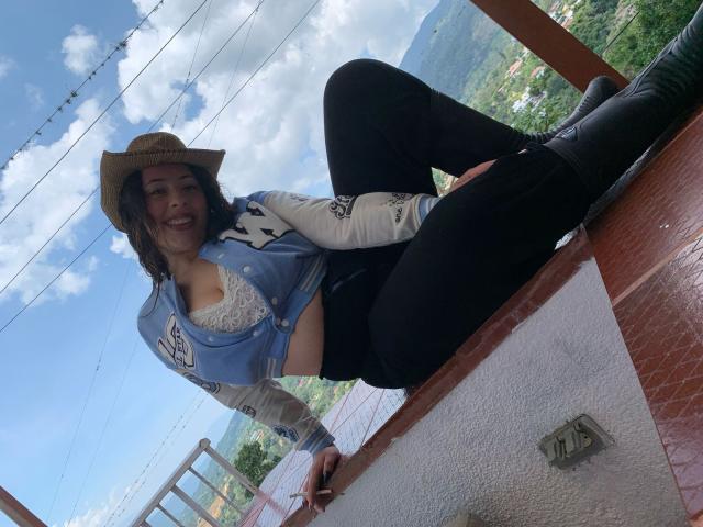 ScarlettParchMusa, in a cowboy hat, blue jacket, black pants, and boots, sits on a red-tiled roof, smiling, with her tits slightly visible, and her pussy not visible.