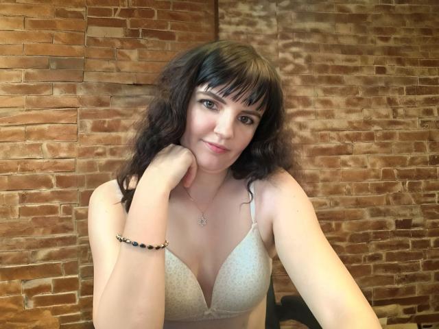 AlicexB with curly black hair, wearing a white bra, has a slight smile, hand on cheek, bracelet on wrist, and a necklace, sitting in front of a brick wall.