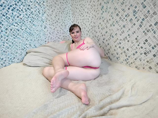 AlicexB with pink lingerie, large ass, and pregnant belly, lying on a bed with white textured blanket, feet in the air, looking at camera, wearing a playful smile.