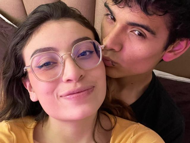 CamilleLiam with round glasses, pink-tinted eyes, and a subtle smile, being gently kissed on the nose by Liam with curly black hair and a black shirt.